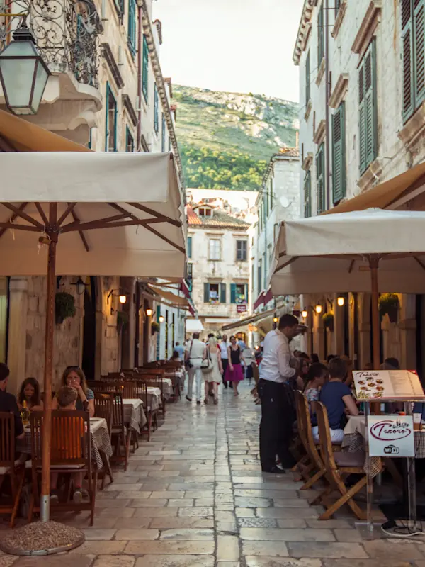 Narrow cobblestone street in Mediterranean town with outdoor cafes, umbrellas, and diners beneath historic buildings and mountain views.