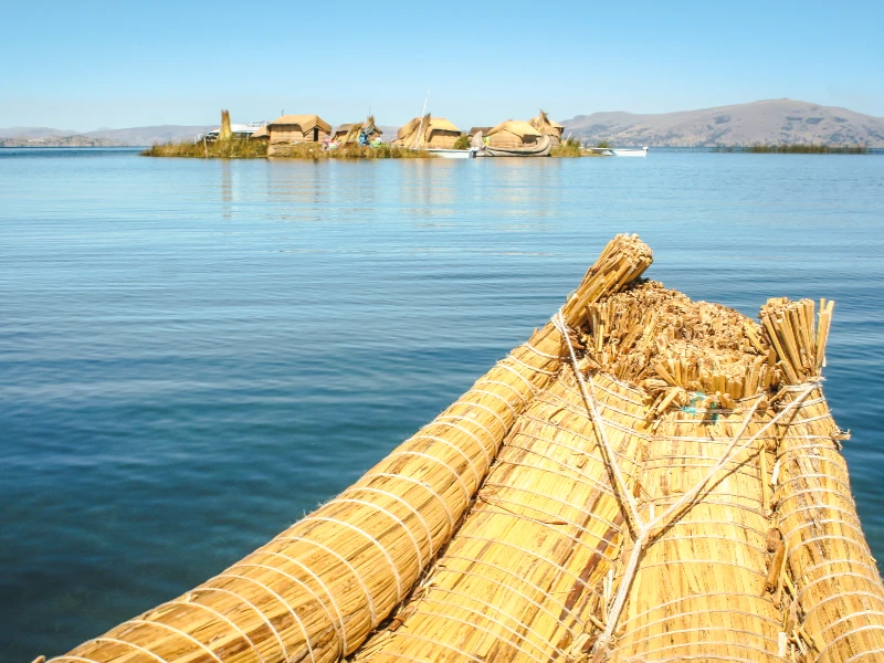 Traditional reed boat on Lake Titicaca approaching floating islands made of totora reeds, with mountains in background.