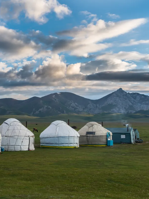 Traditional white yurts on a green plain with dramatic mountains in the background under a cloudy blue sky.
