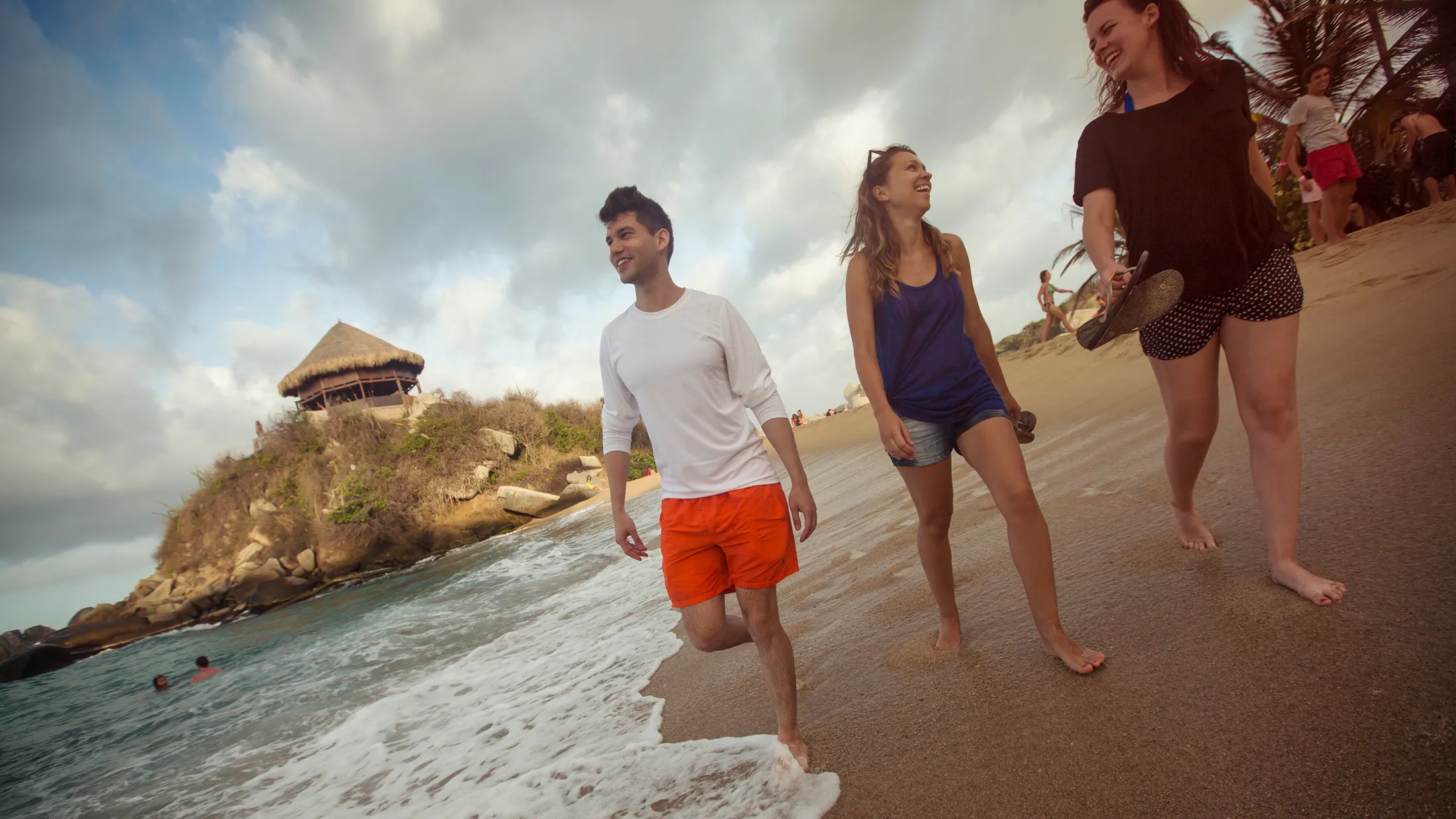 Three friends laughing and walking barefoot along a sandy beach, with a thatched hut on a rocky hill in the background.