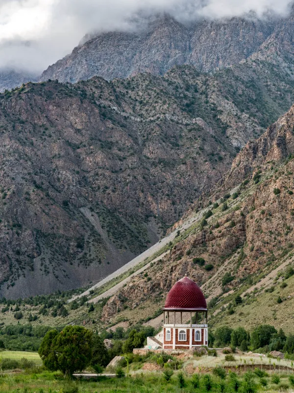 Small domed building with red roof nestled at the base of dramatic rocky mountains partially shrouded in clouds.