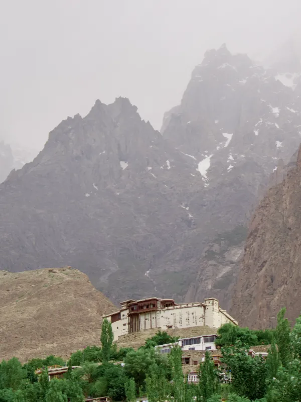 A white monastery nestled among green trees at the base of towering, misty mountain peaks with patches of snow.