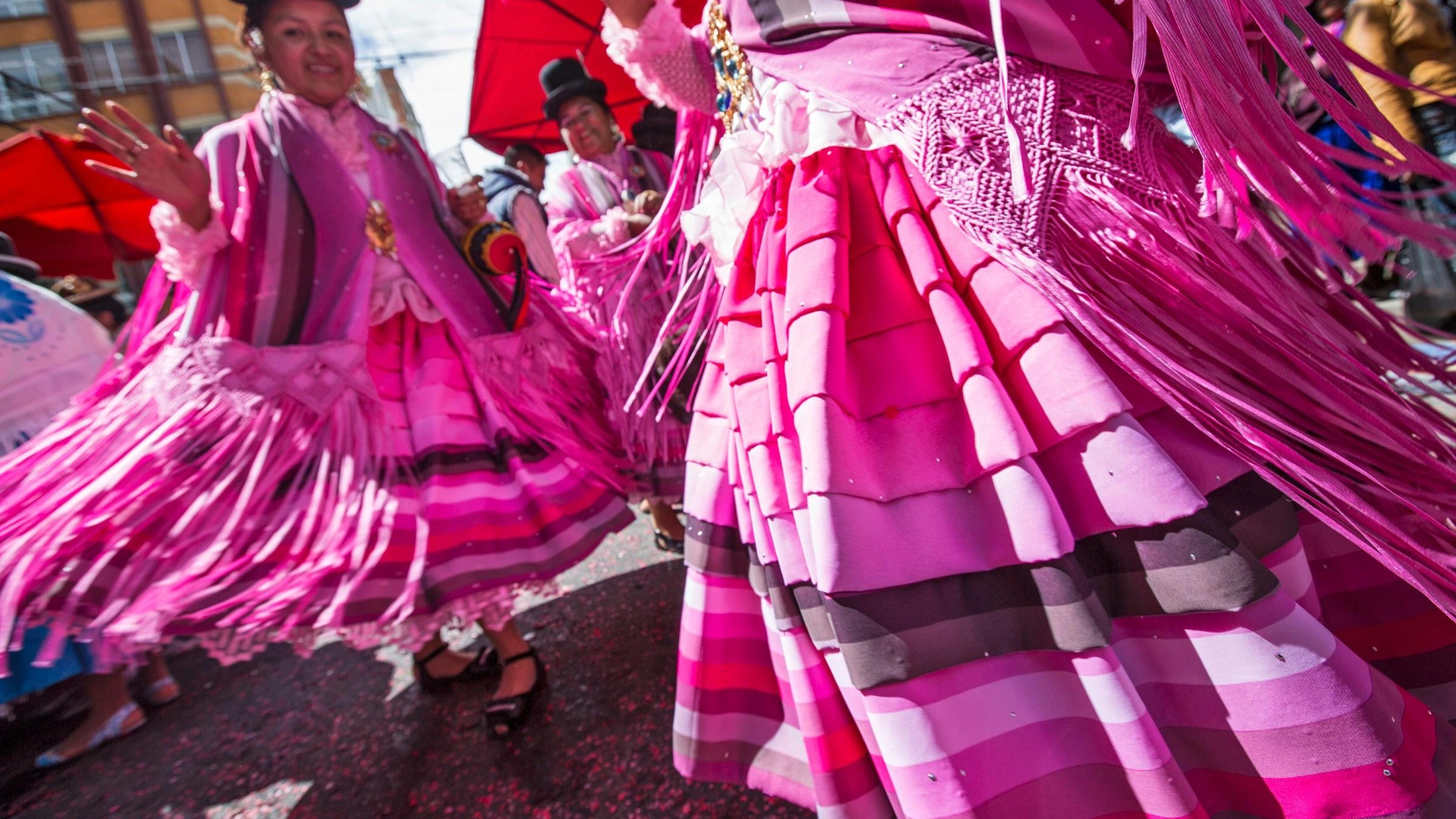 Women in vibrant pink fringed traditional dresses dance at a colorful Latin American street festival parade.