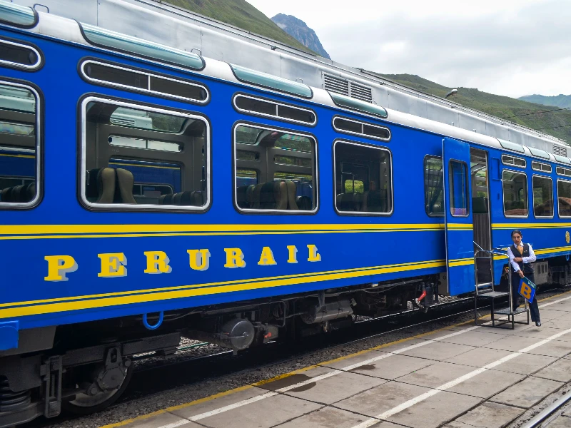 Blue and yellow PeruRail train car stopped at a station platform with mountains in the background.