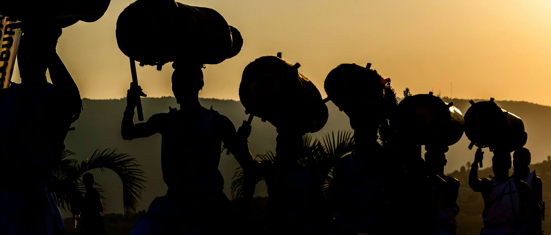 Silhouettes of people carrying large round objects on their heads against a golden sunset sky, with palm trees visible in background.
