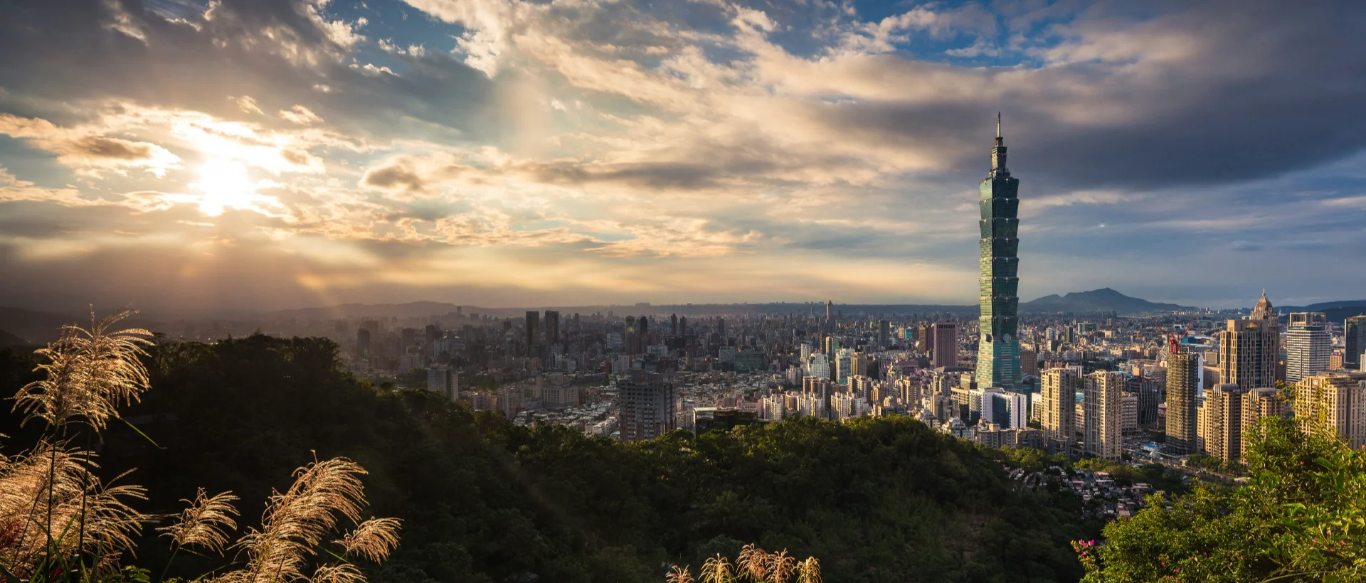 Panoramic view of Taipei skyline at sunset with Taipei 101 tower rising above the city, viewed from a forested hillside.