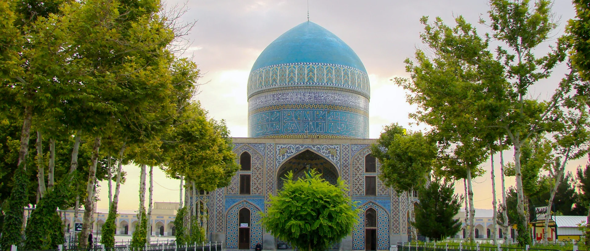 Turquoise-domed mosque with intricate blue tilework surrounded by green trees against a cloudy sky.