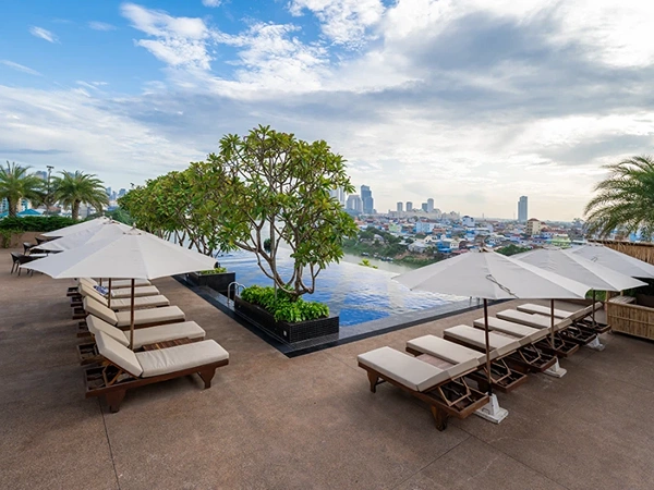 Rooftop infinity pool with lounge chairs, white umbrellas, a tree centerpiece, and a city skyline backdrop under a cloudy sky.