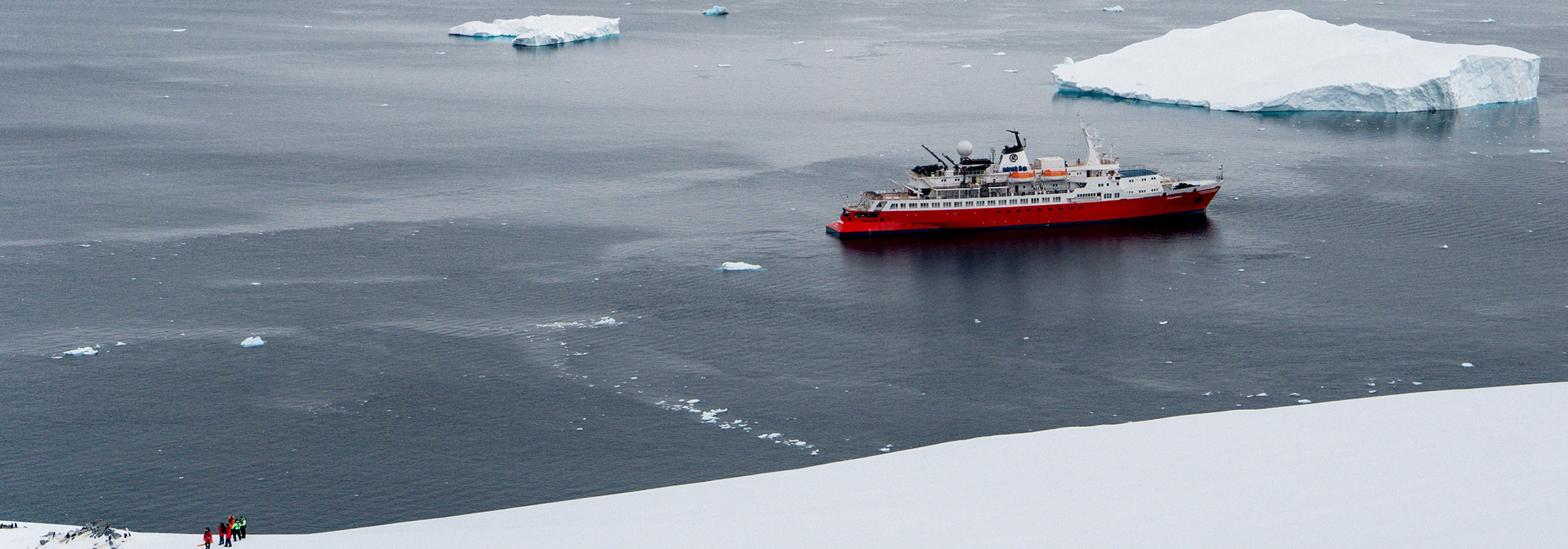 A red and white expedition ship sails calm Antarctic waters, surrounded by snow-covered mountains and glaciers under a blue sky.