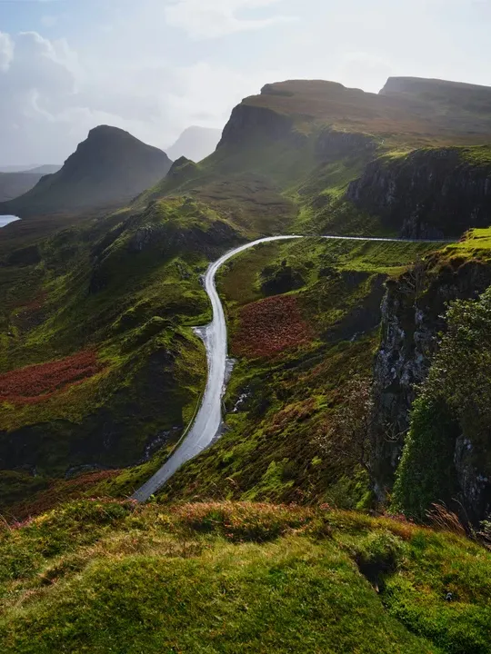 Winding road cutting through rugged green mountains in Scotland, with dramatic cliffs and misty peaks in the distance.