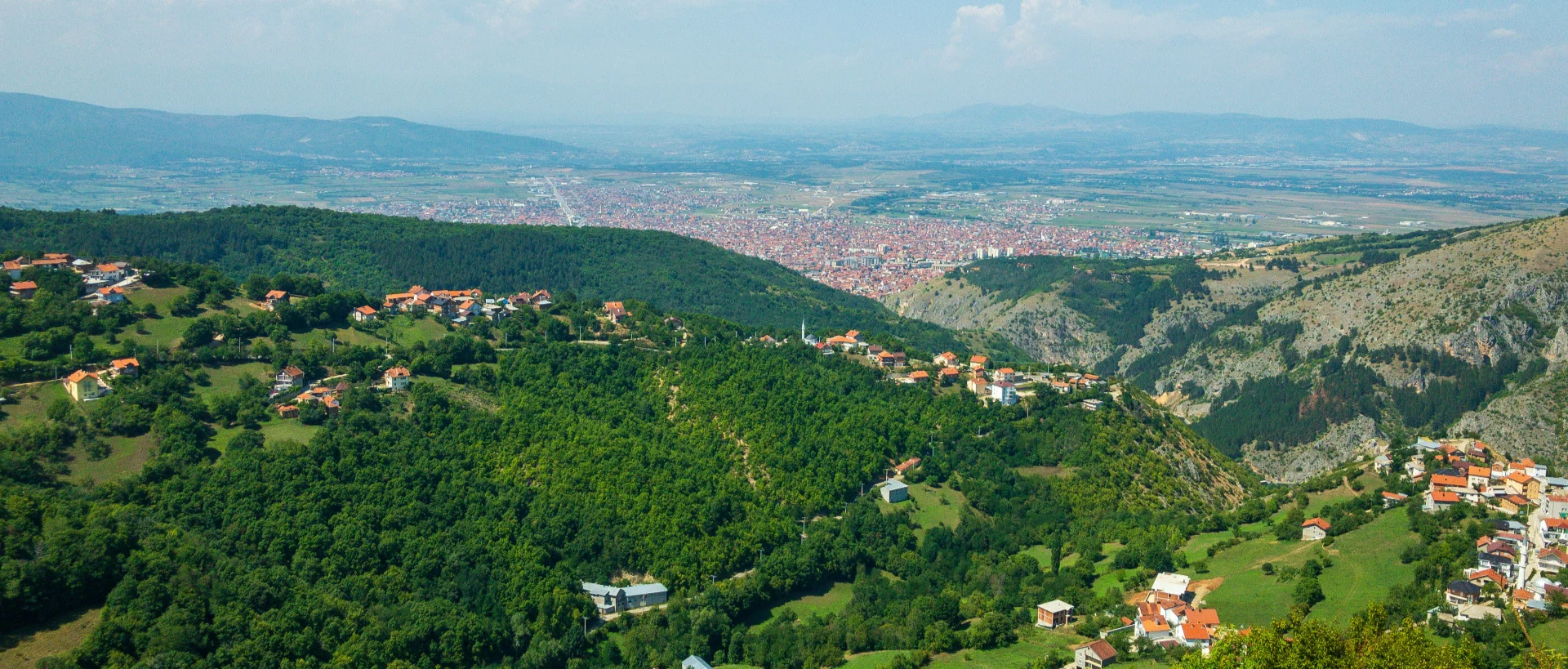Aerial view of mountain villages with red-roofed houses nestled in green hills, overlooking a large city in a valley with mountains beyond.