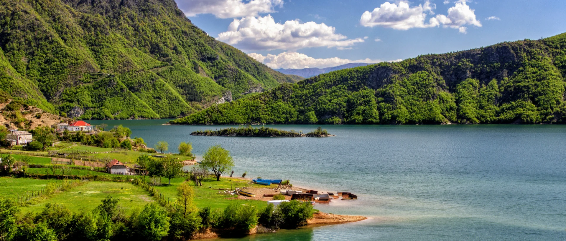 Serene mountain lake surrounded by lush green hills, with small houses along the shore and a tiny island in the blue water.