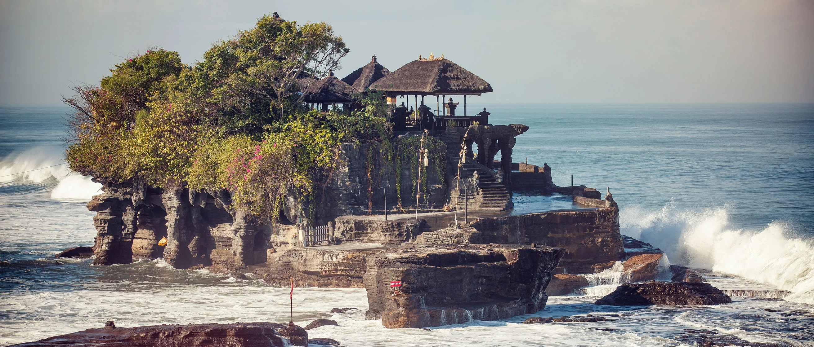 Tanah Lot temple perched on rocky outcrop in Bali, surrounded by ocean waves with thatched pavilions and lush vegetation.