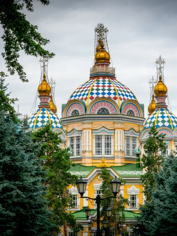 Colorful Orthodox cathedral with blue checkered domes topped with golden spires, framed by evergreen trees against a cloudy sky.