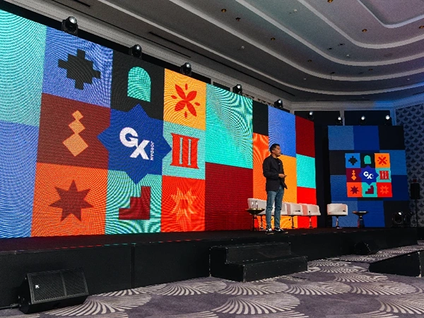 A speaker stands on a colorful conference stage with a large LED backdrop displaying the GX logo in vibrant orange, teal, and blue panels.