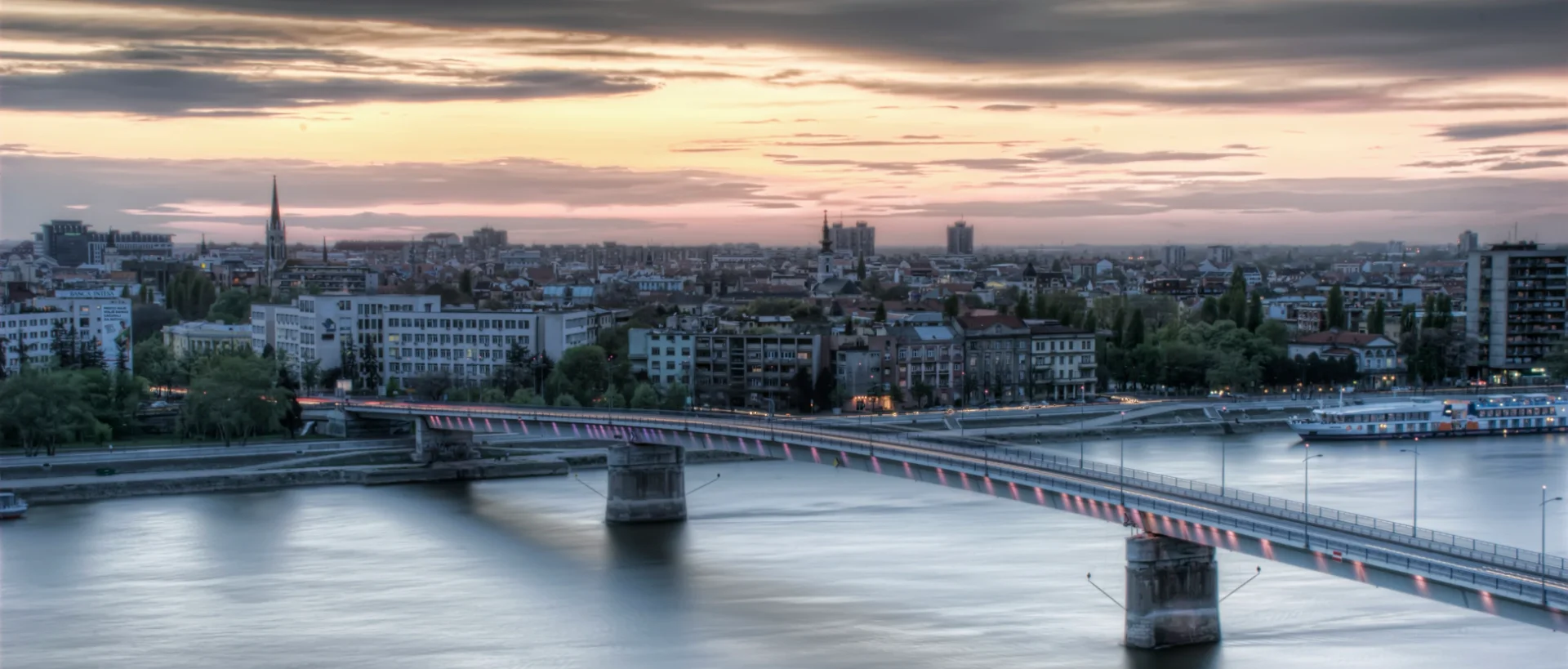 Panoramic view of a European city skyline at dusk with a bridge spanning across a river, illuminated by soft pink lights.