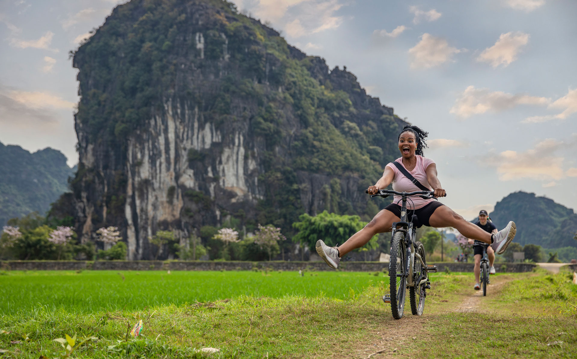 A joyful woman rides a bike with legs outstretched near green rice fields, with dramatic karst mountains in the background.