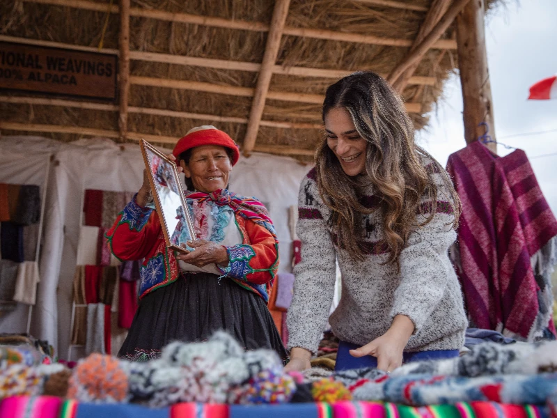 Two women in a thatched market stall with traditional textiles; one in colorful Peruvian dress showing handcrafted items to a visitor.