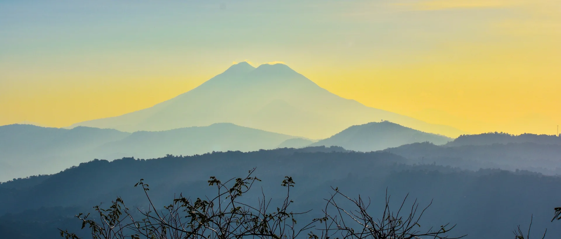 Silhouette of a mountain with twin peaks against a golden sunset sky, with layers of blue hills in the foreground.