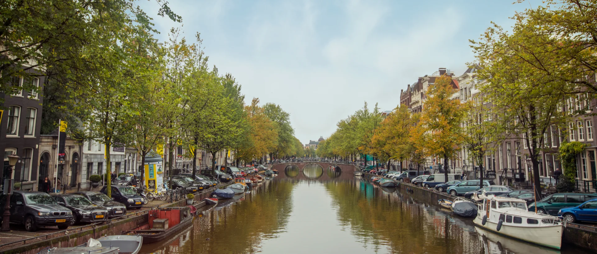 Amsterdam canal lined with autumn trees, historic buildings, parked cars, and boats on both sides, with a bridge in the distance.