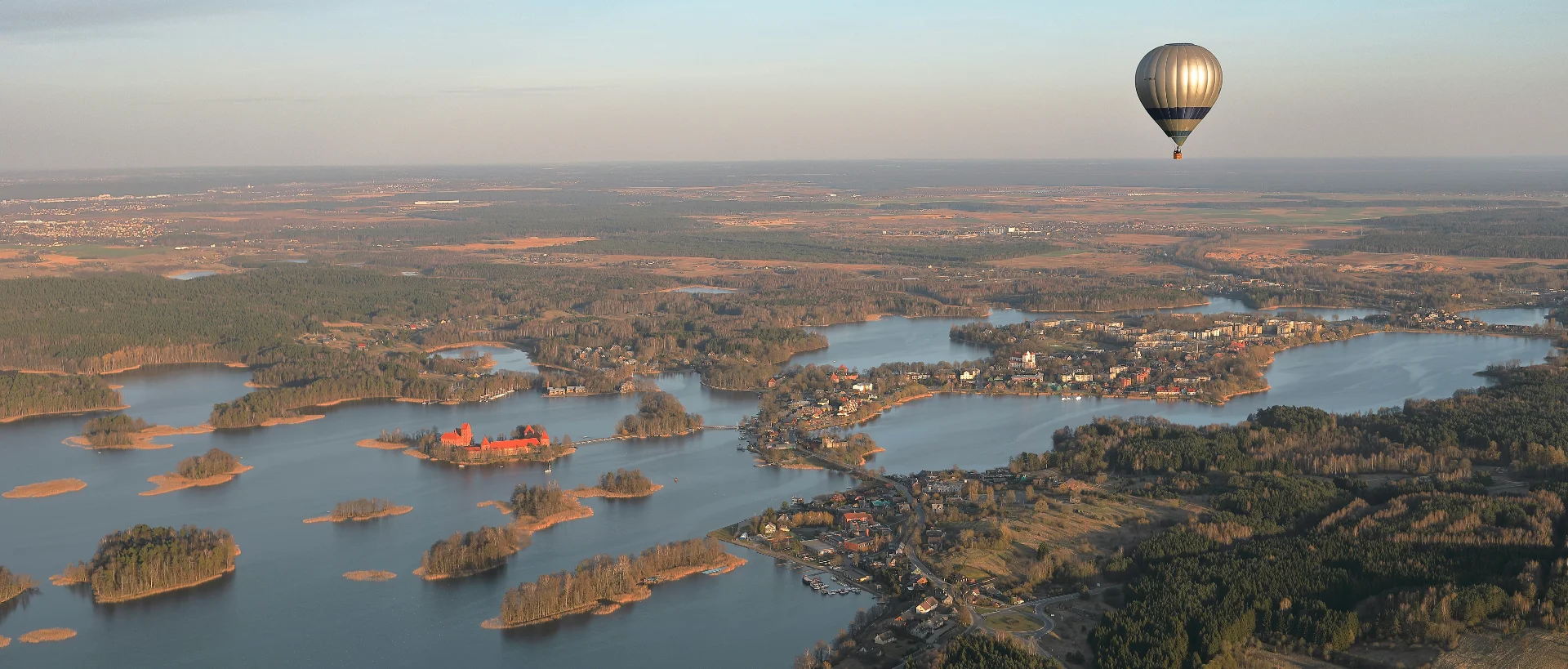 Silver hot air balloon floating over a scenic landscape with blue lakes, small islands, forests, and scattered settlements below.