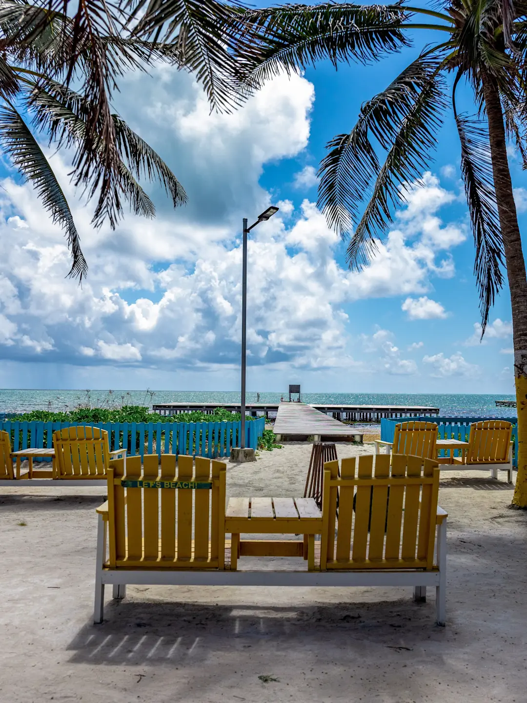 Yellow beach chairs facing a wooden pier extending into turquoise waters, framed by palm trees under a bright blue sky.