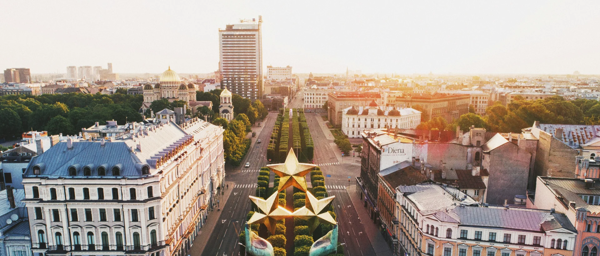 Aerial view of Riga cityscape at sunset, featuring historic buildings, a boulevard with golden star monuments, and an Orthodox cathedral.