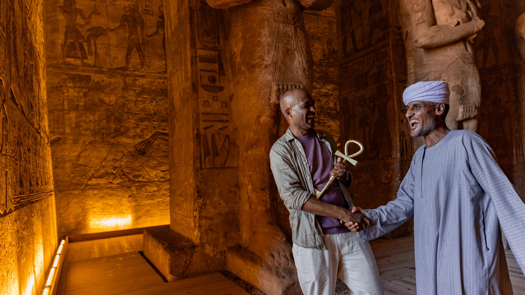 Two men laugh and shake hands inside an ancient Egyptian temple with hieroglyph-covered walls, one holding a golden ankh.