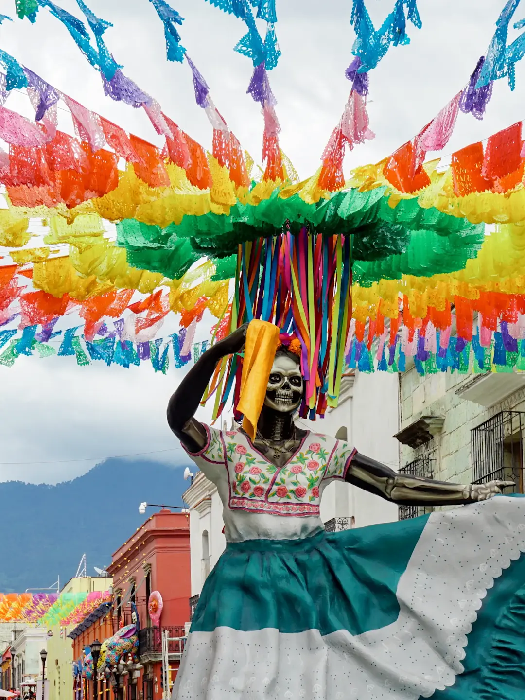 Colorful Day of the Dead skeleton figure in traditional dress beneath vibrant paper decorations on a Mexican street.