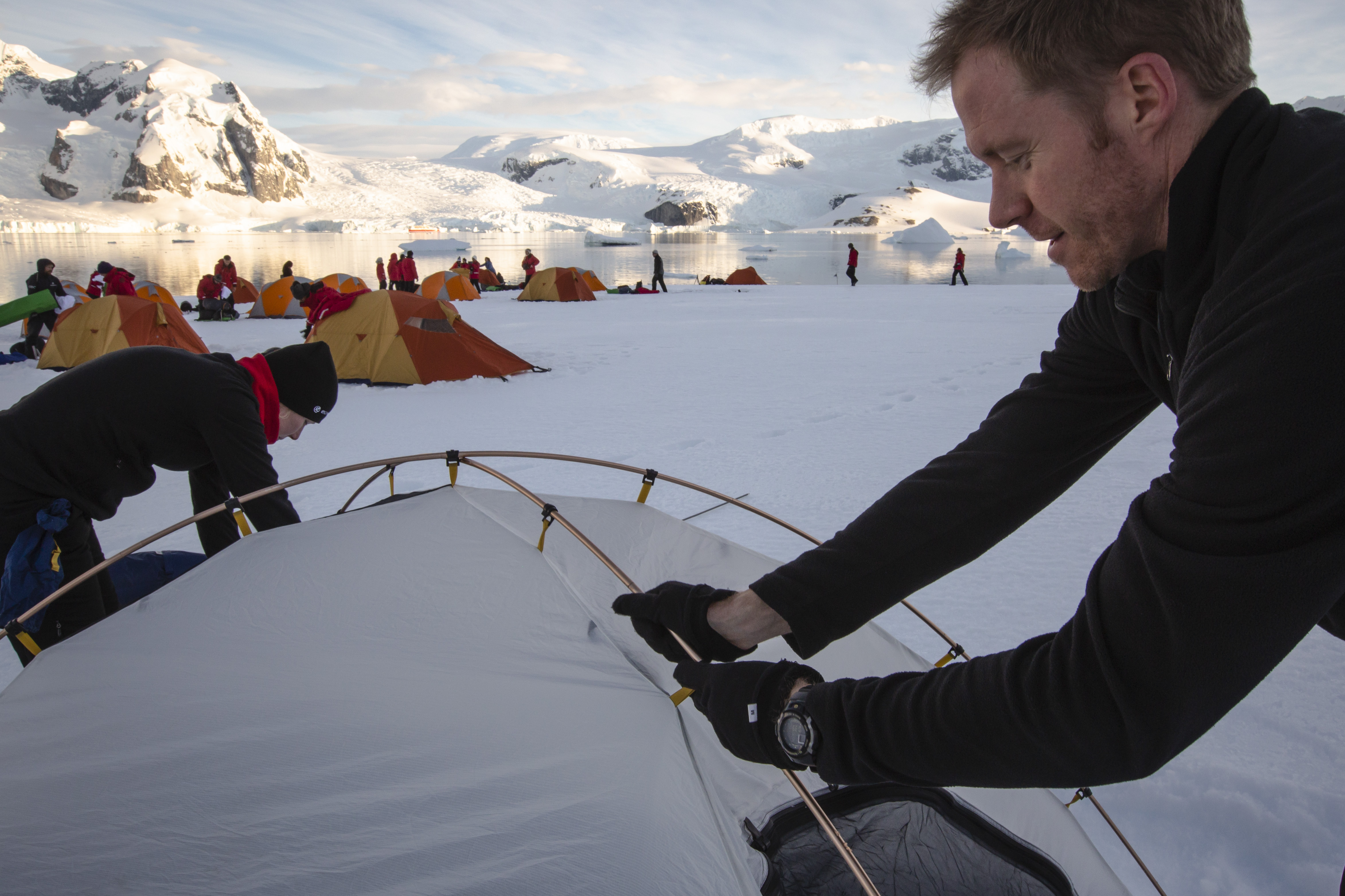 A man sets up a tent on snow in Antarctica, with orange tents, people in red jackets, icy water, and snowy mountains behind.
