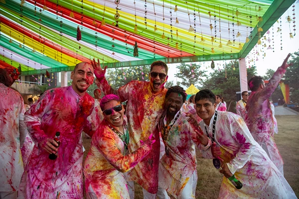 Group of joyful people covered in colorful powder at a Holi festival, posing under vibrant red, green, and yellow decorations.