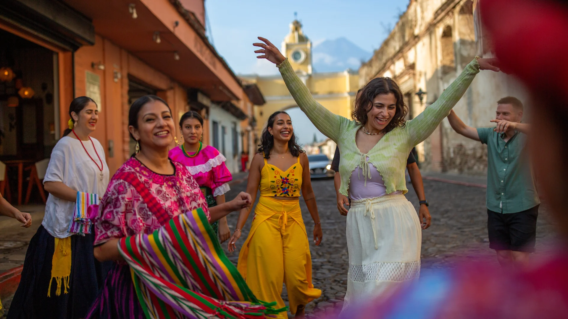 Joyful group in colorful traditional clothing dancing on a cobblestone street in Antigua, Guatemala, with a yellow clock tower visible.