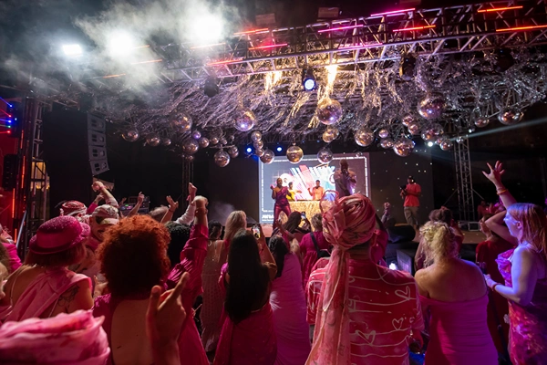 Crowd in pink outfits dancing at a lively concert with disco balls, stage lighting, and performers on a lit stage.