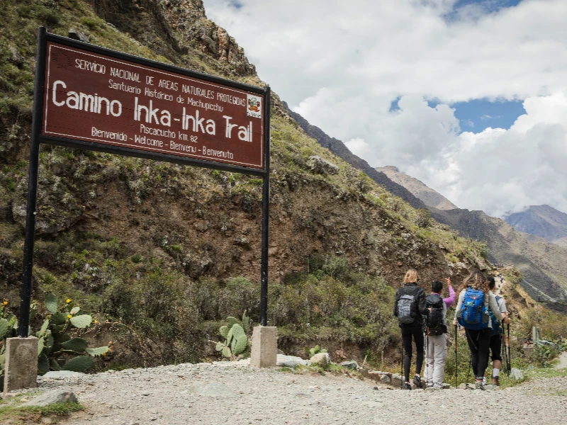 Hikers with backpacks at the entrance sign for Camino Inka-Inka Trail in Peru, surrounded by mountainous landscape.