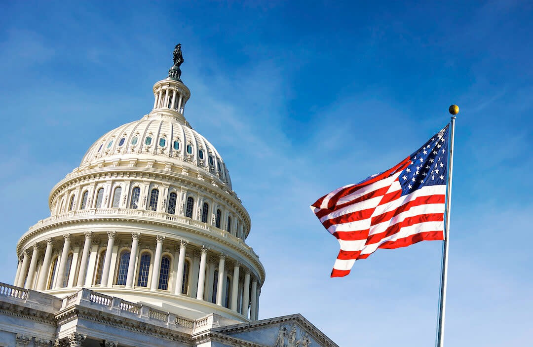 The image shows the United States Capitol building with its iconic dome, set against a clear blue sky. An American flag flies prominently in the foreground, slightly to the right of the Capitol dome, symbolizing recent discussions on unemployment insurance changes and FFCRA support.