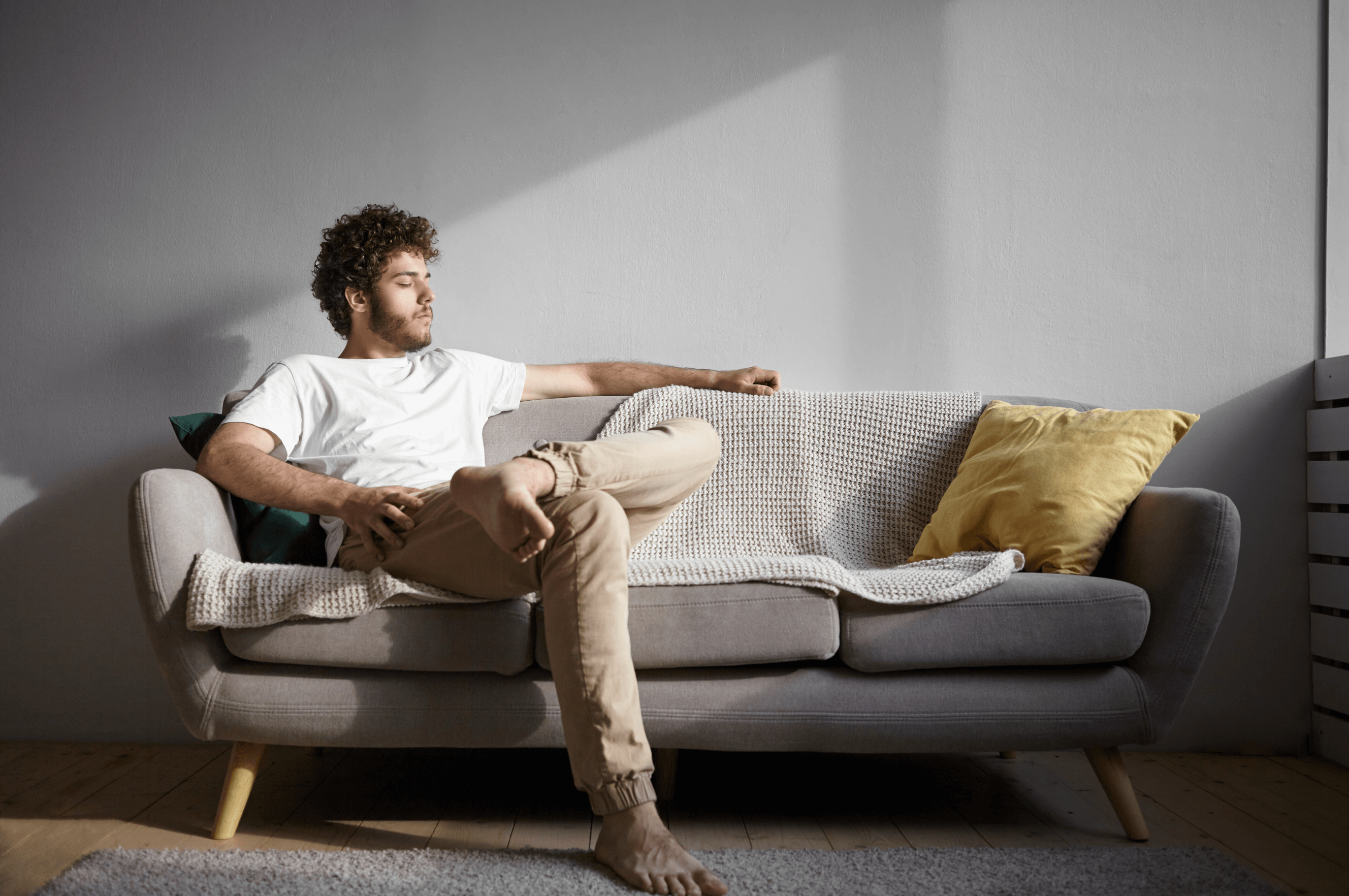 A man with curly hair is sitting on a gray sofa, leaning back casually with one arm resting on the back. He wears a white t-shirt and beige pants. Sunlight streams in, casting shadows behind him as he ponders cost-effective leave solutions. A yellow pillow is beside him.
