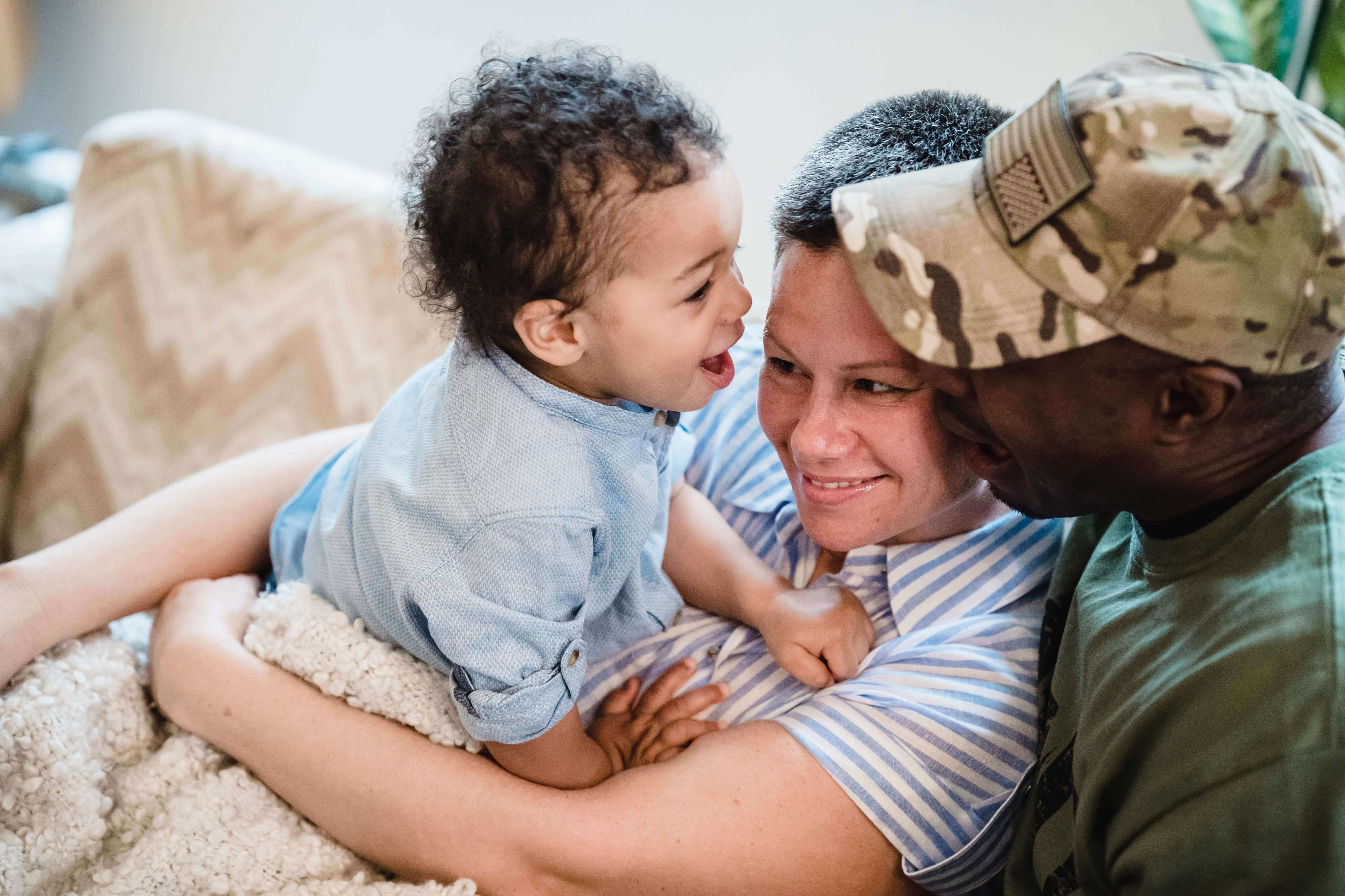 A happy family of three, including a young child, playfully interacting on a couch. The child, wearing a blue shirt, is laughing and being held by an adult in a striped shirt, while a person in a camouflage cap—likely benefiting from Military Leave Pay Protection—smiles warmly at them.