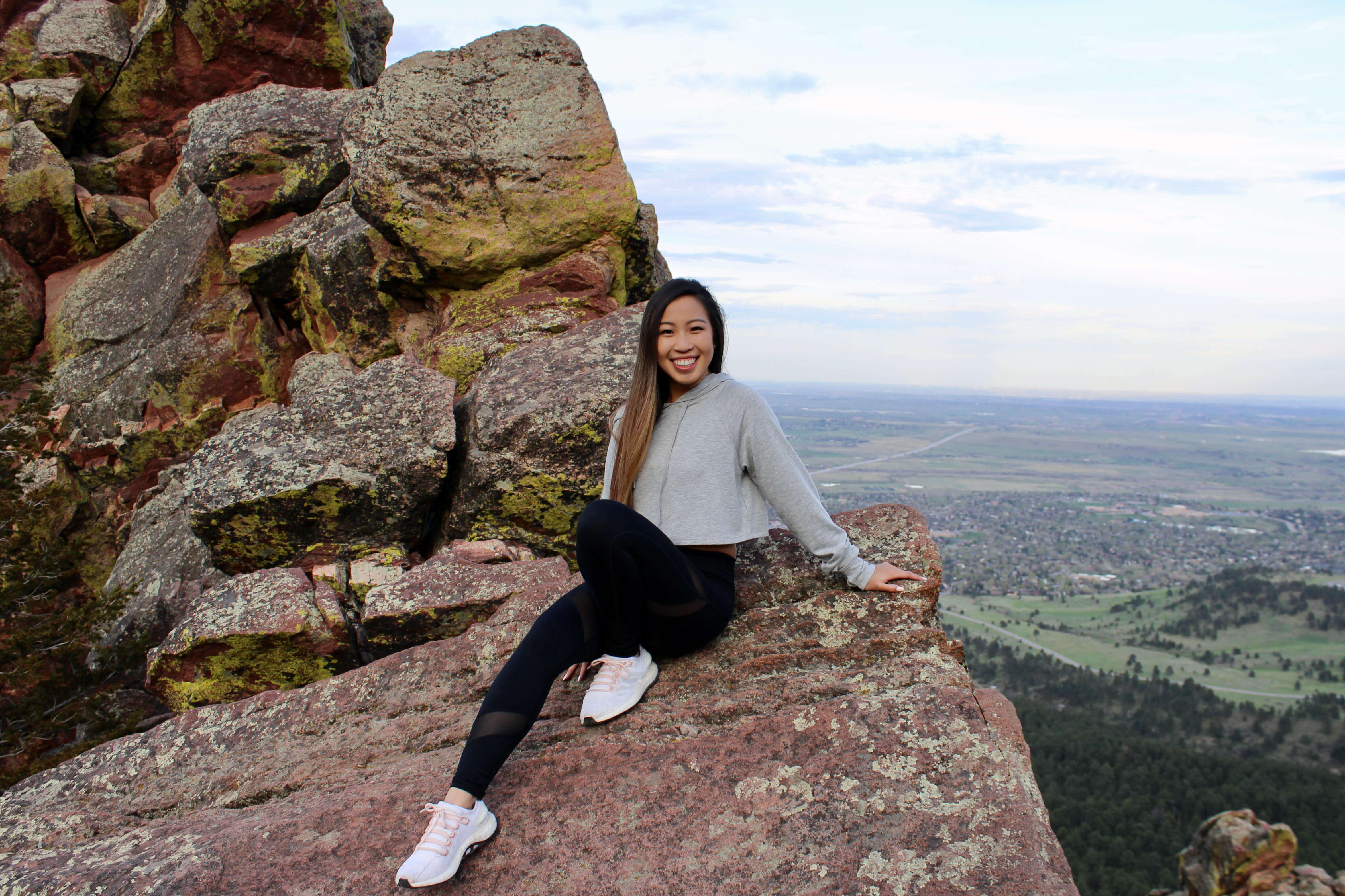 A person (Kelly) with long hair sits on a large, rugged rock formation, smiling towards the camera. They are wearing a gray sweatshirt, black leggings, and white sneakers. Behind them is a scenic view of a distant town, lush greenery, and a cloudy sky.
