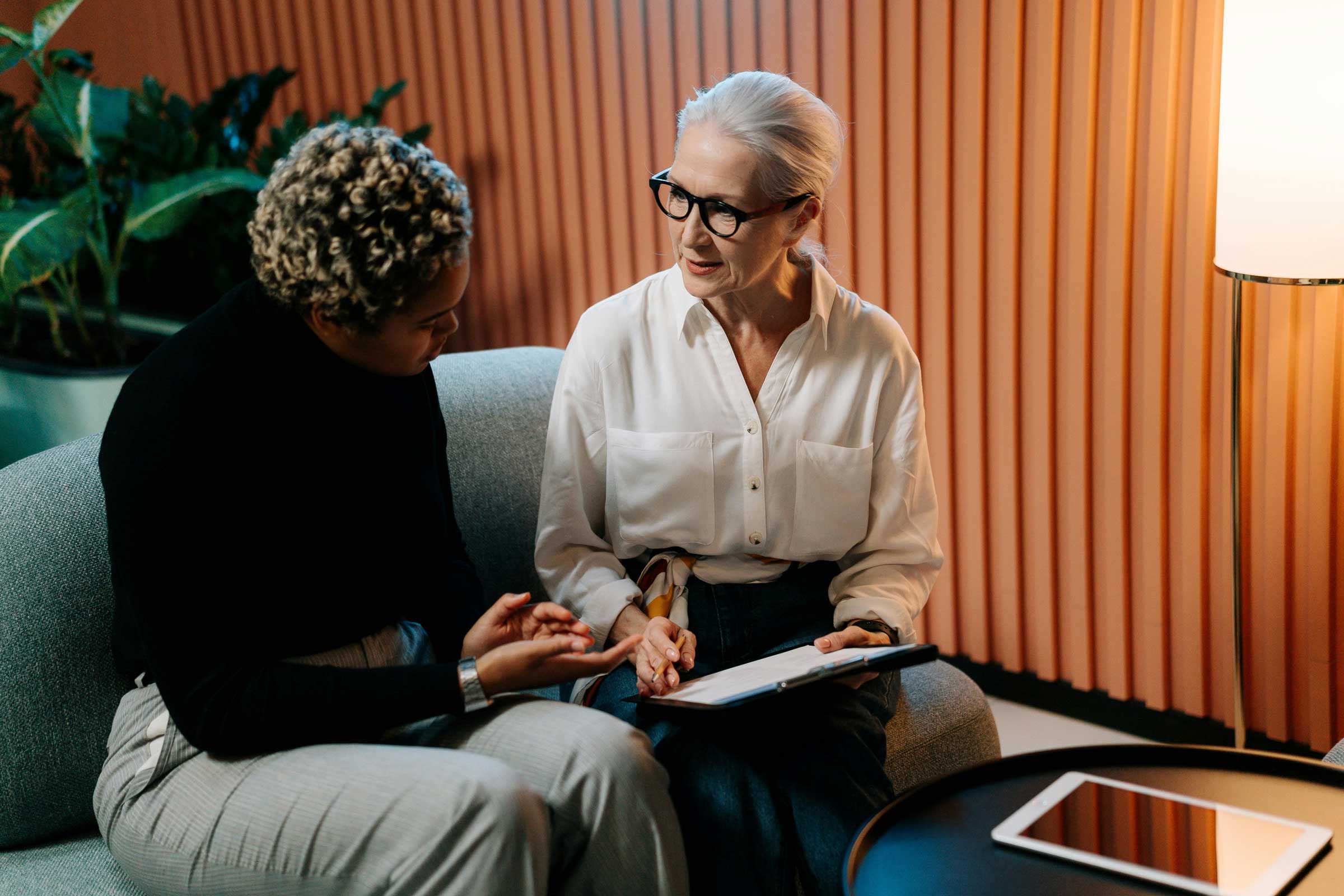Two women are seated on a sofa engaged in a conversation about employee rights. One is holding a tablet, and they are in a room with plants and a corrugated wall. A round table and lamp are nearby, adding to the cozy setting of their discussion on employer responsibilities.
