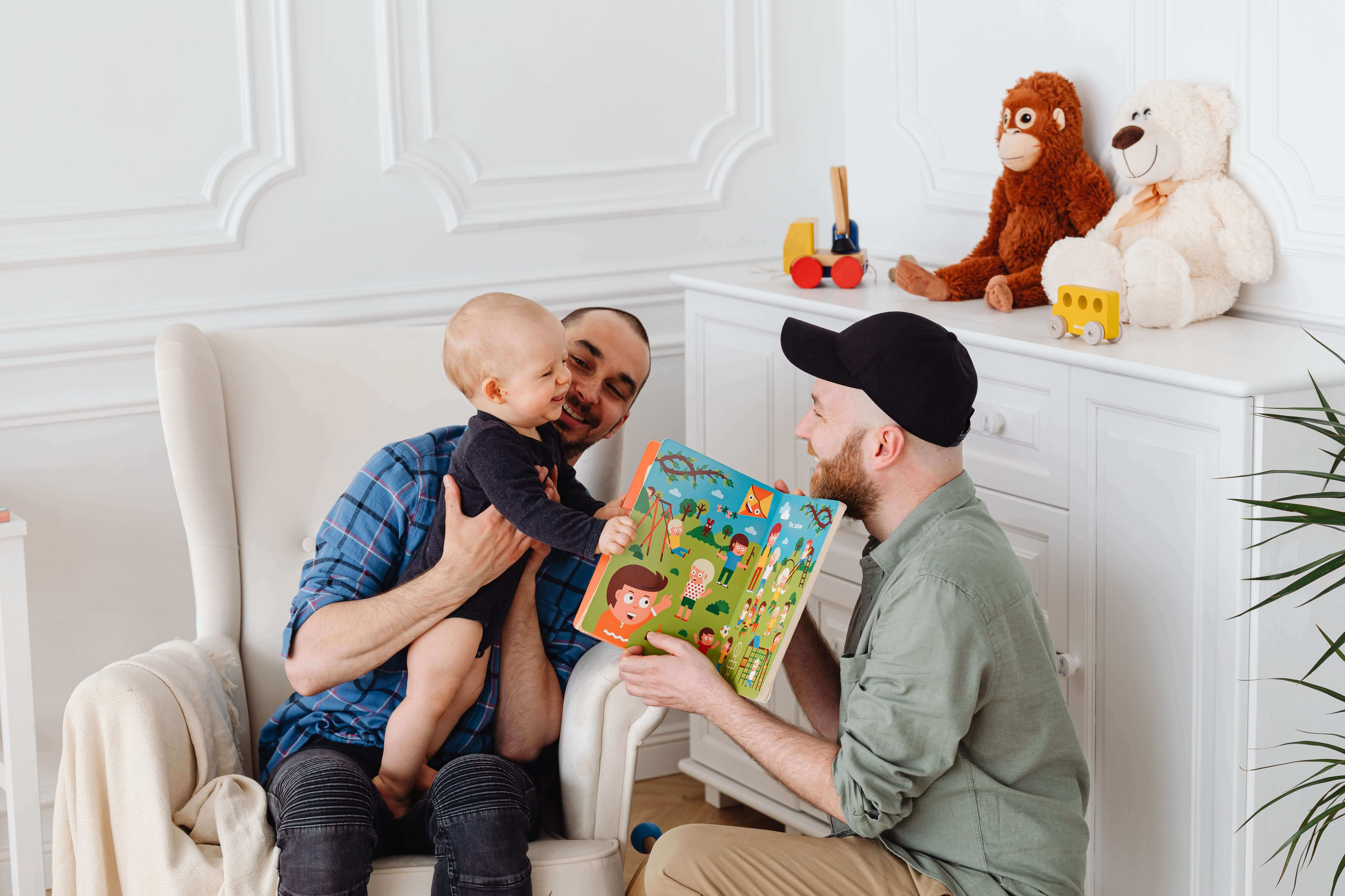 A baby sits on one man's lap while another man kneels in front of them holding an open colorful children's book. They are indoors, with toy animals and other children's items visible on a nearby dresser. All three are smiling and appear to be enjoying the moment, thanks to paid time off allowing quality family bonding.