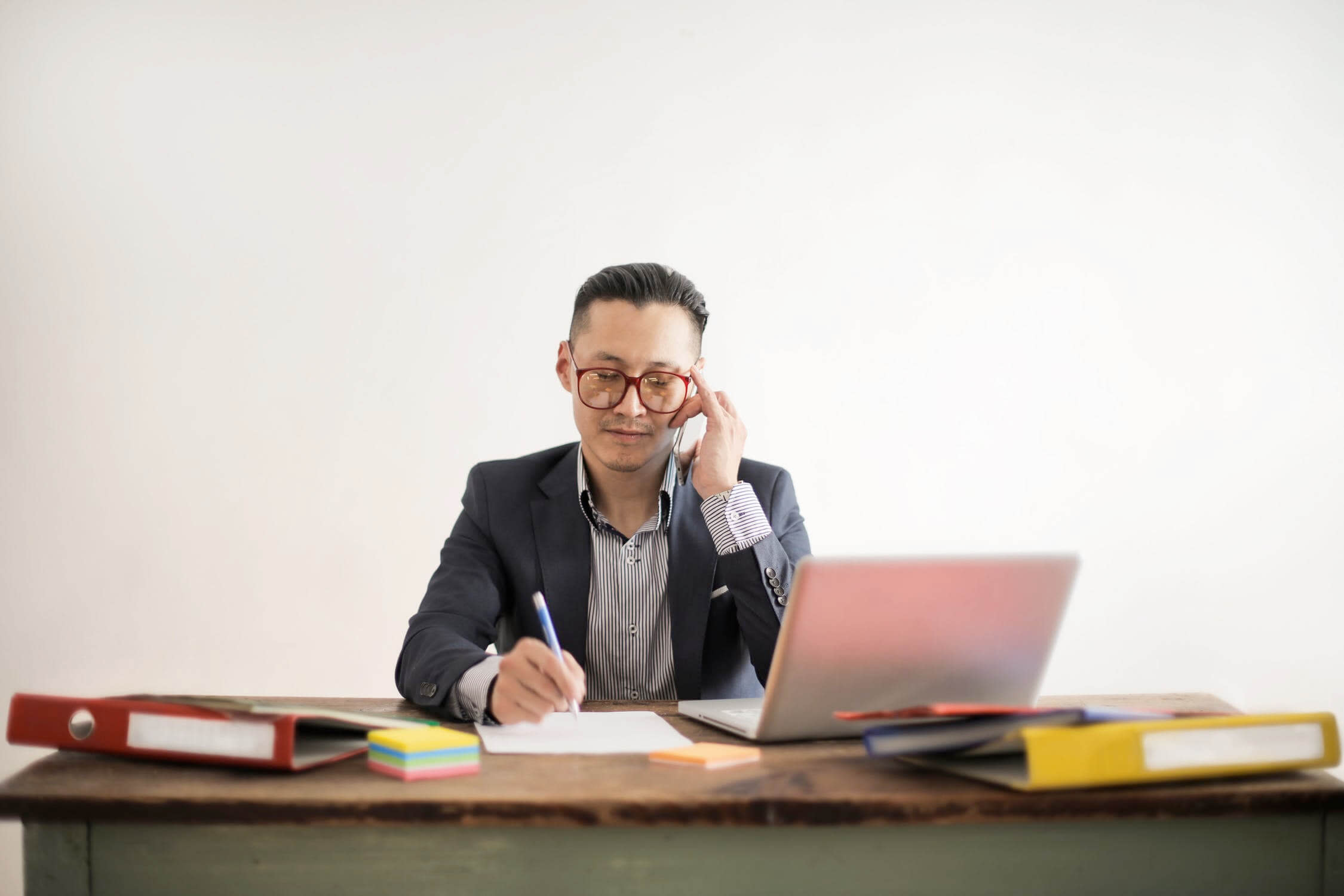 A person wearing glasses and business attire sits at a wooden desk, talking on the phone about disability benefits questions while writing on a piece of paper. An open laptop, colorful folders, and sticky notes are also on the desk. The background is neutral.