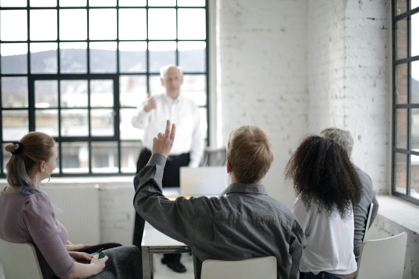A group of four people sits around a table in a bright room, with large windows and mountains visible outside. A person in a white shirt stands at the front speaking about FFCRA compliance, while one individual raises their hand, signaling participation.
