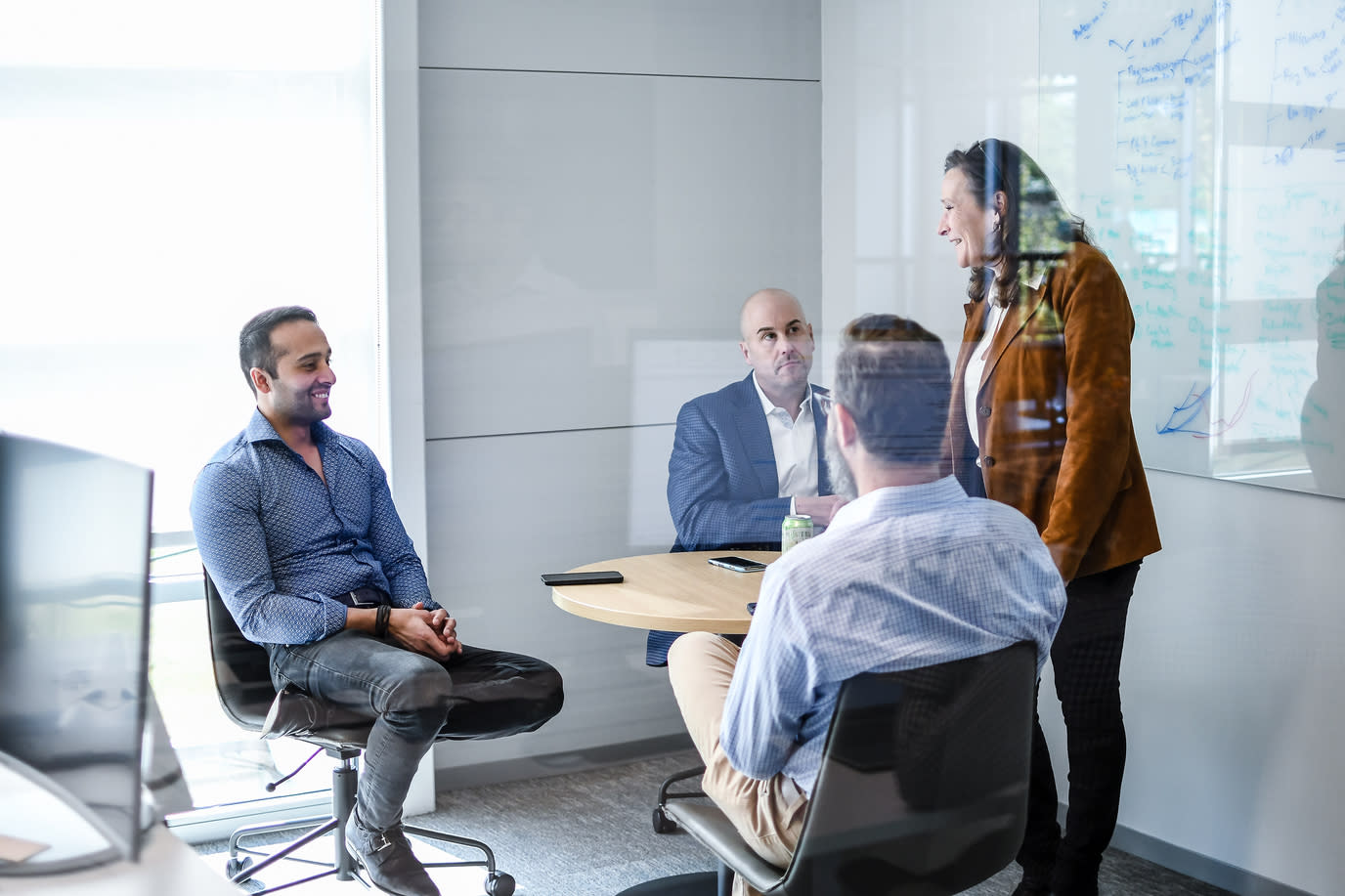 Four people are having a meeting in a glass-walled office. Three individuals are seated around a small circular table, while one person stands, addressing the group. A whiteboard with notes on the Cart.com case study is visible in the background. Everyone appears engaged in conversation.