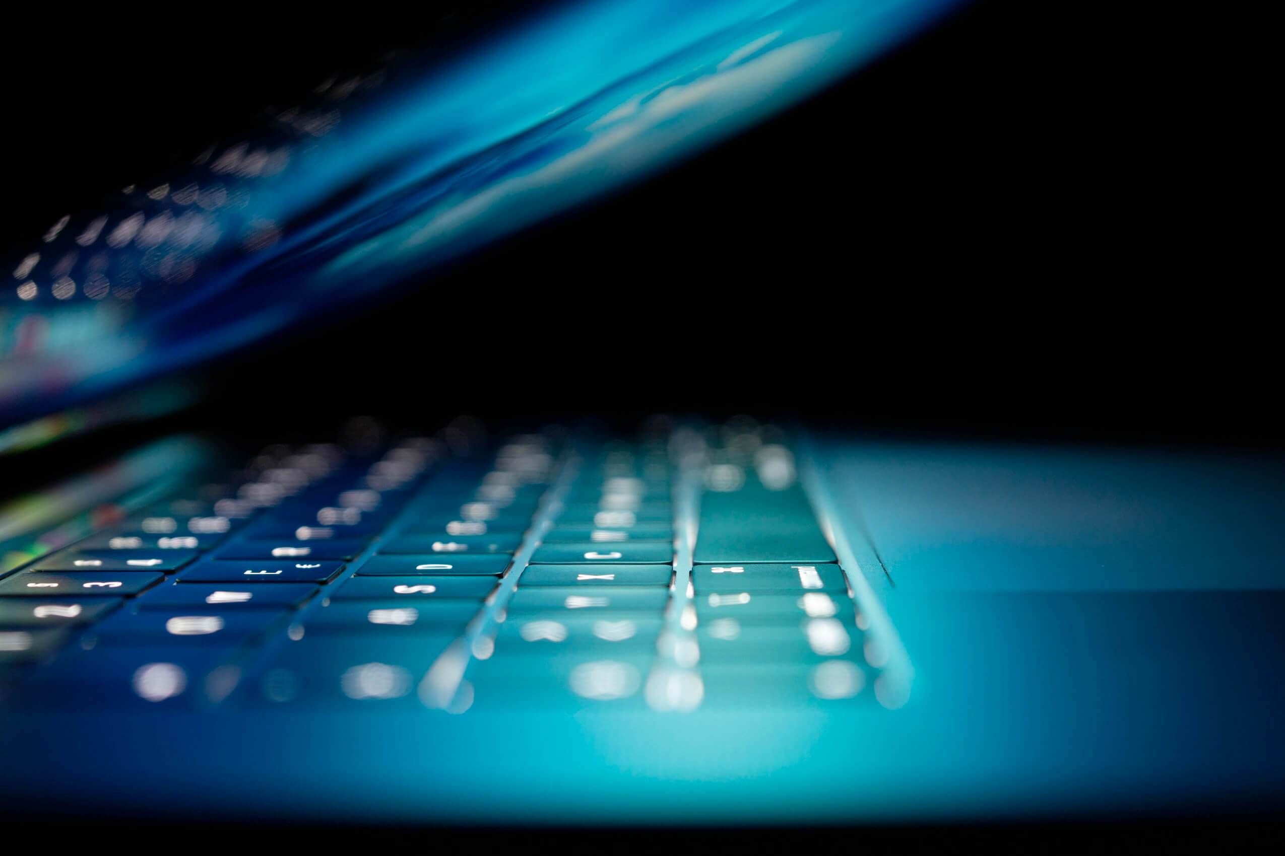 A close-up photo of a laptop keyboard with the screen partially closed. The keys, illuminated with a blue light, create a sleek and futuristic atmosphere, reflecting SaaS security standards. The focus is sharp on the keys in the foreground, while the background fades into darkness.
