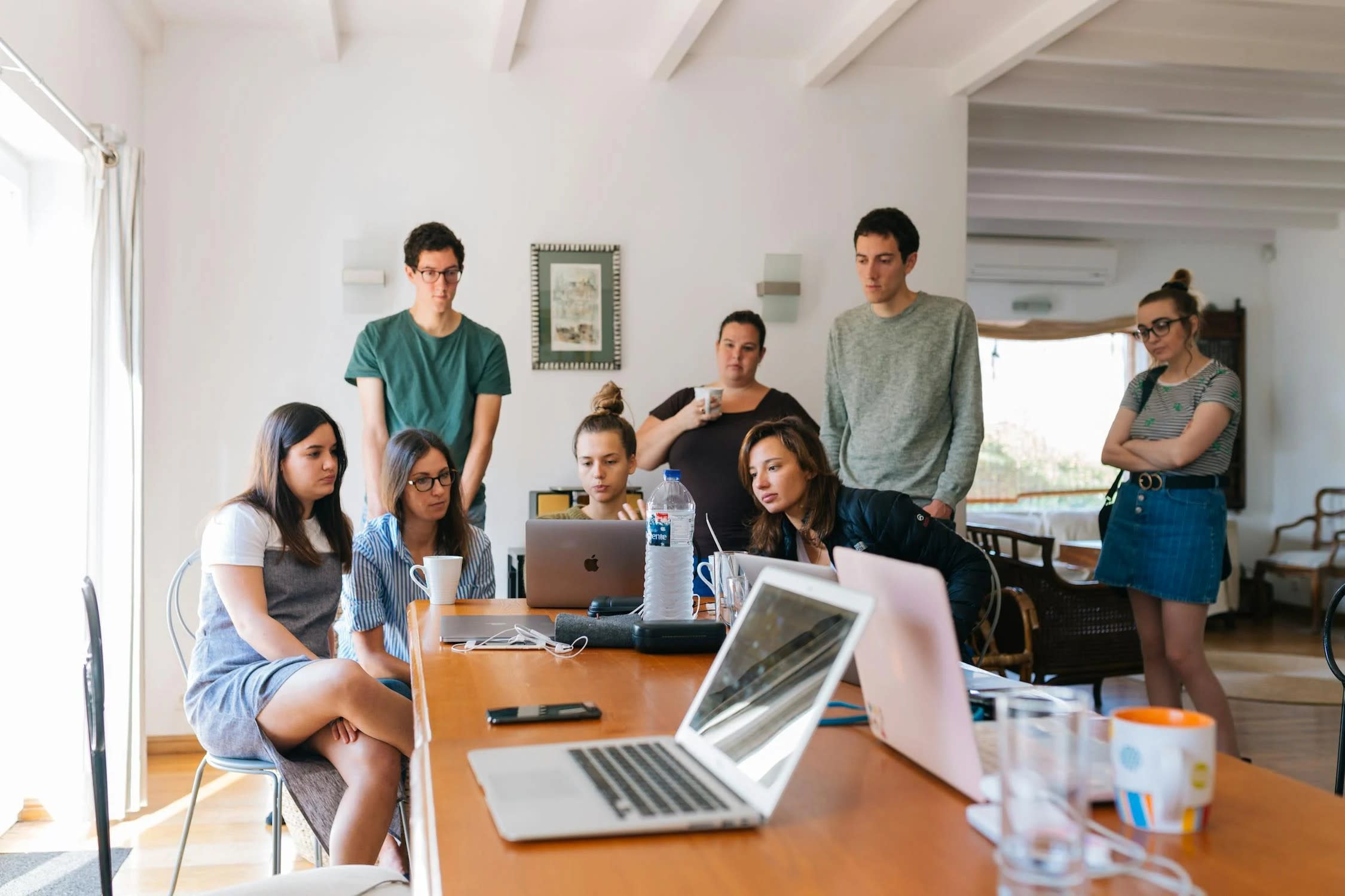 A group of people gathered around a table with laptops, immersed in the Sparrow case study discussion. Several are seated, while others stand behind, observing. The room is well-lit with natural light streaming through a window on the left.