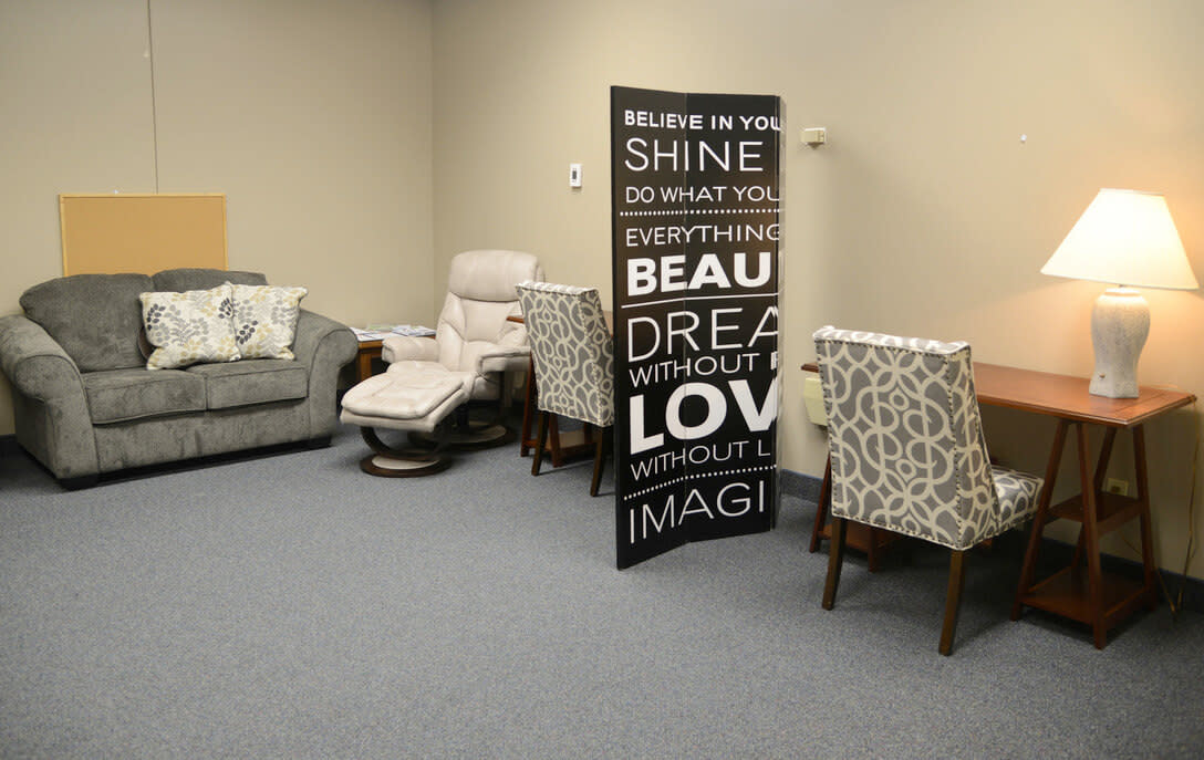 A cozy, affordable lactation room setup with a grey couch decorated with patterned cushions, a beige reclining chair, a wooden divider with inspirational words, and two patterned chairs at wooden desks. A table lamp sits on one of the desks, emitting a warm light.