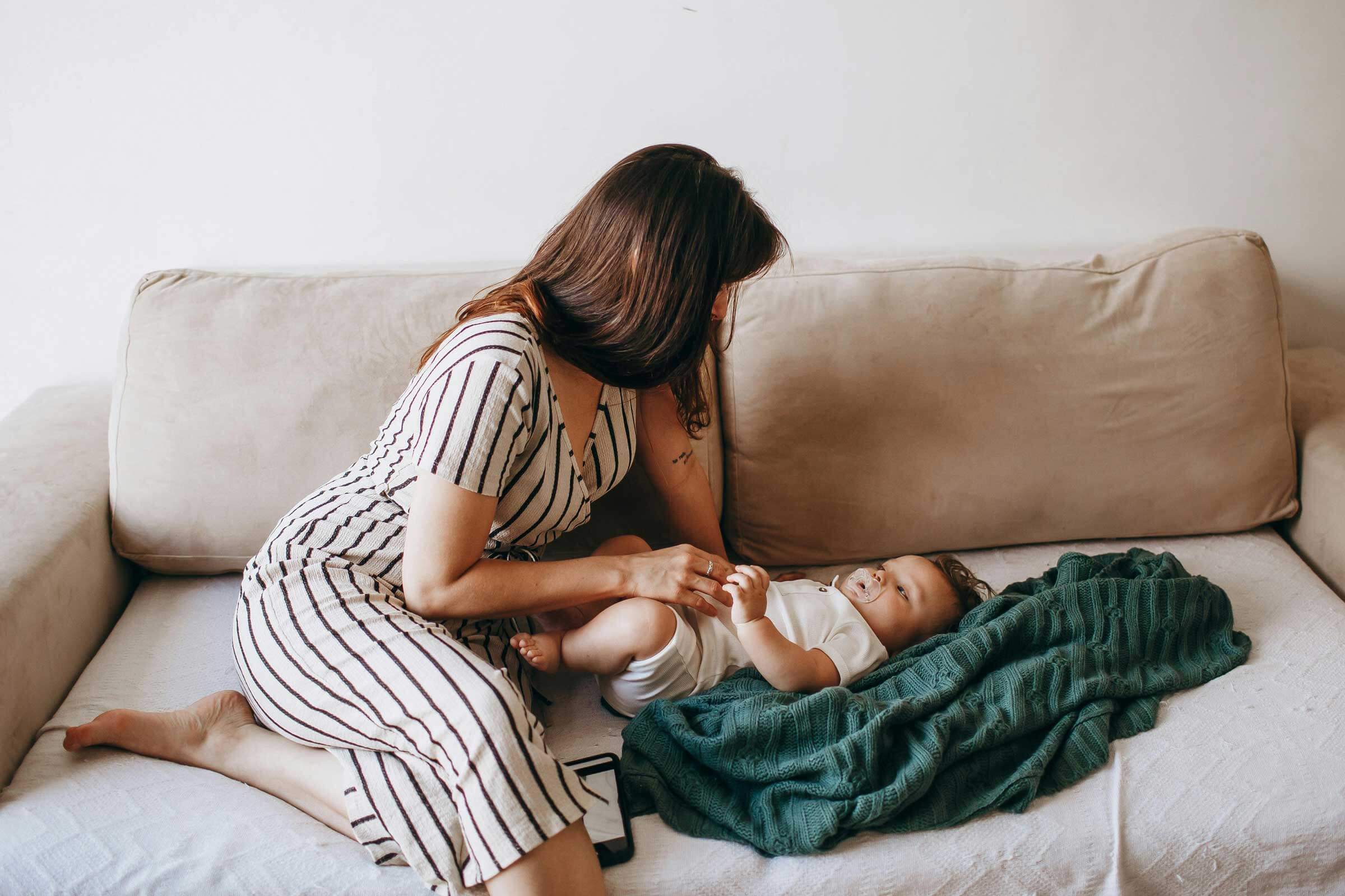 A woman in a striped dress kneels on a beige couch, gently holding the baby's hand. The baby, snug on a white and green blanket, wears a white onesie. This serene scene emanates the cozy charm of Rhode Island.