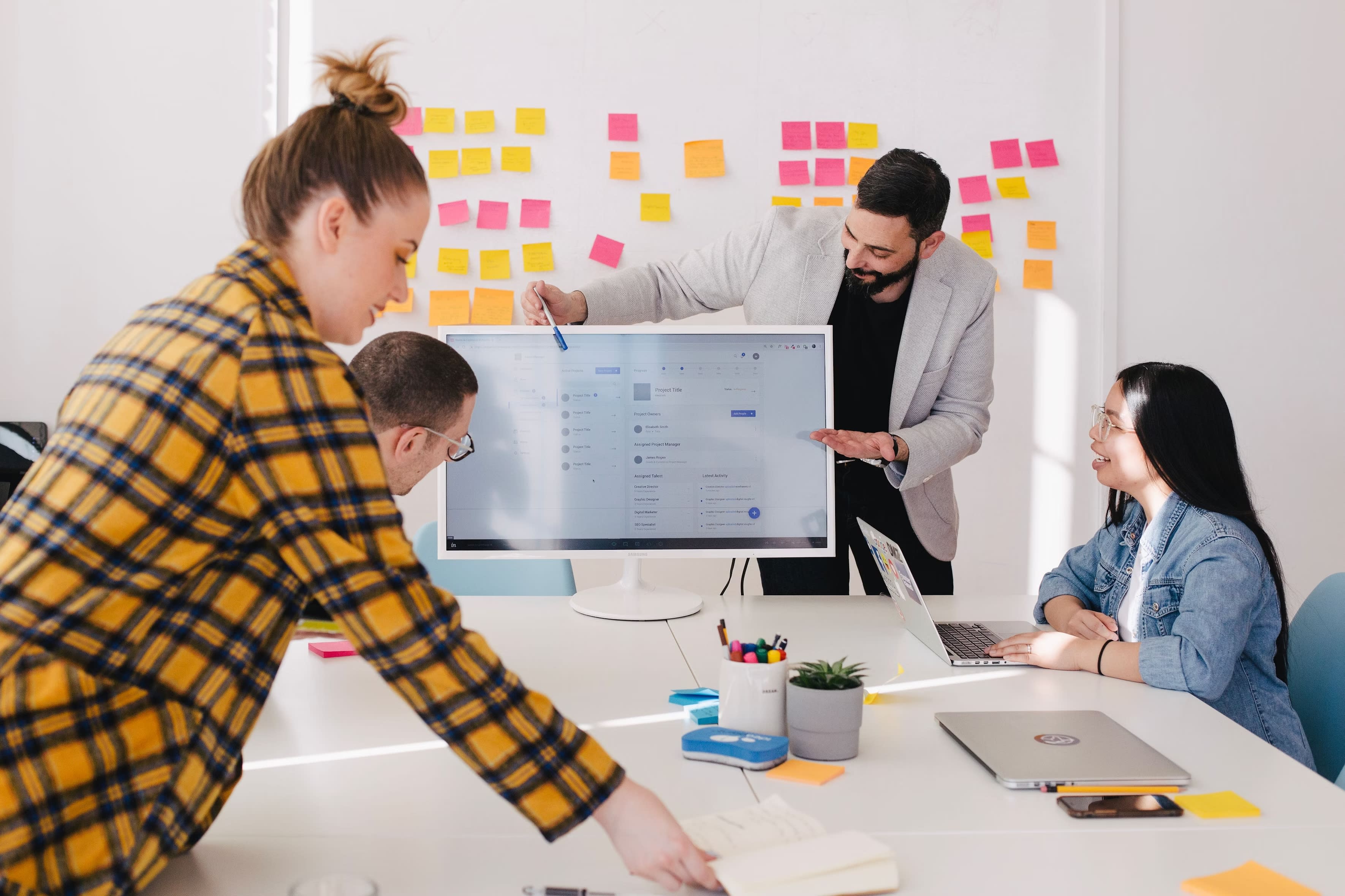 A diverse group of four people in a bright office is engaged in a meeting on benefits administration. One person points at a computer screen displaying a website, while colorful sticky notes adorn the wall. Laptops and office supplies are spread across the table.