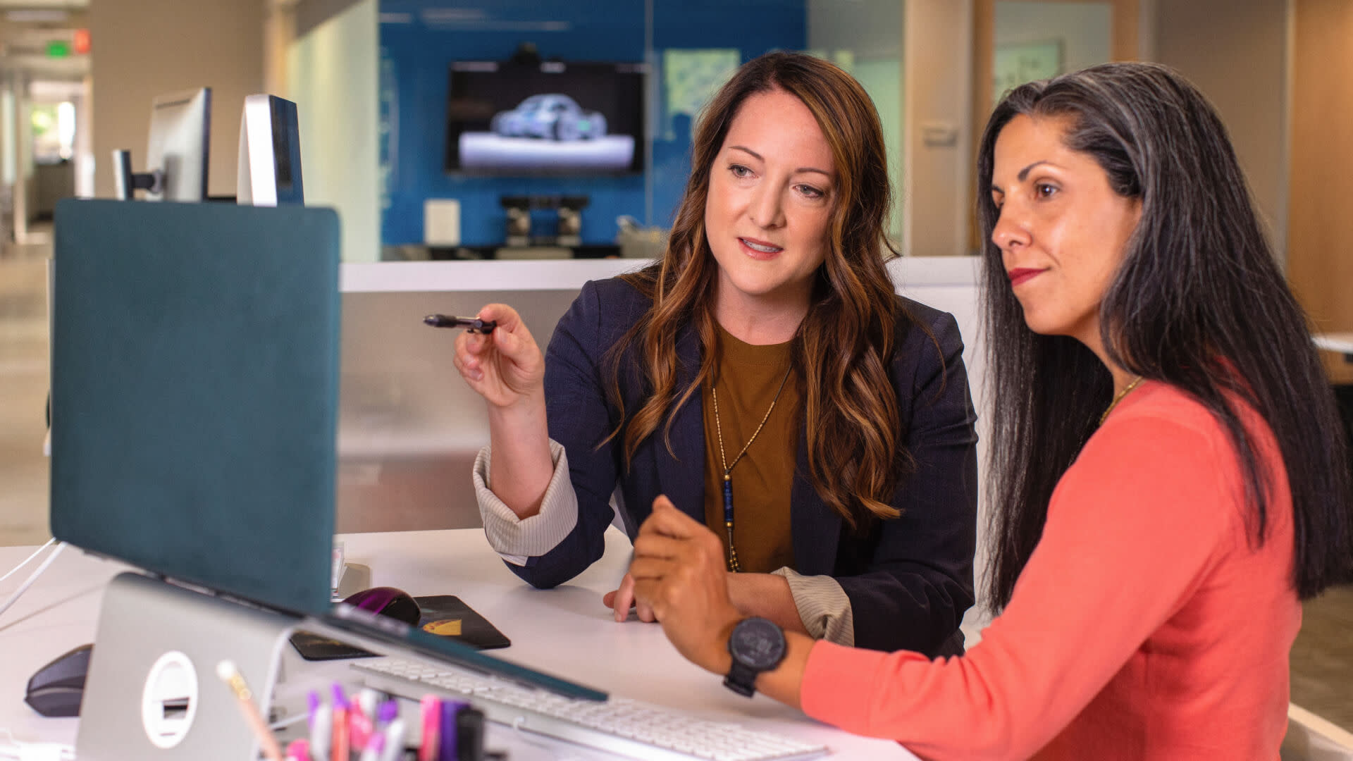 Two women sit at a desk in an office, looking at a computer screen. One woman, with long brown hair, is pointing at the screen while explaining something about operational efficiency. The other woman, with long black hair, listens attentively. Both appear focused and engaged in the discussion.