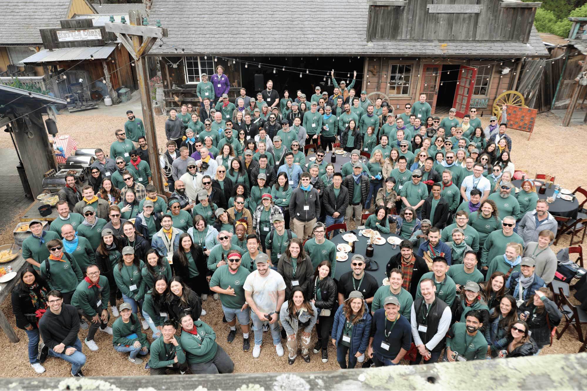 A large group of people, many wearing green hoodies, gather for a group photo outside at a rustic venue with wooden buildings. They smile and wave at the camera, showcasing their camaraderie. There are tables with food and drinks around them. Some people stand while others sit.